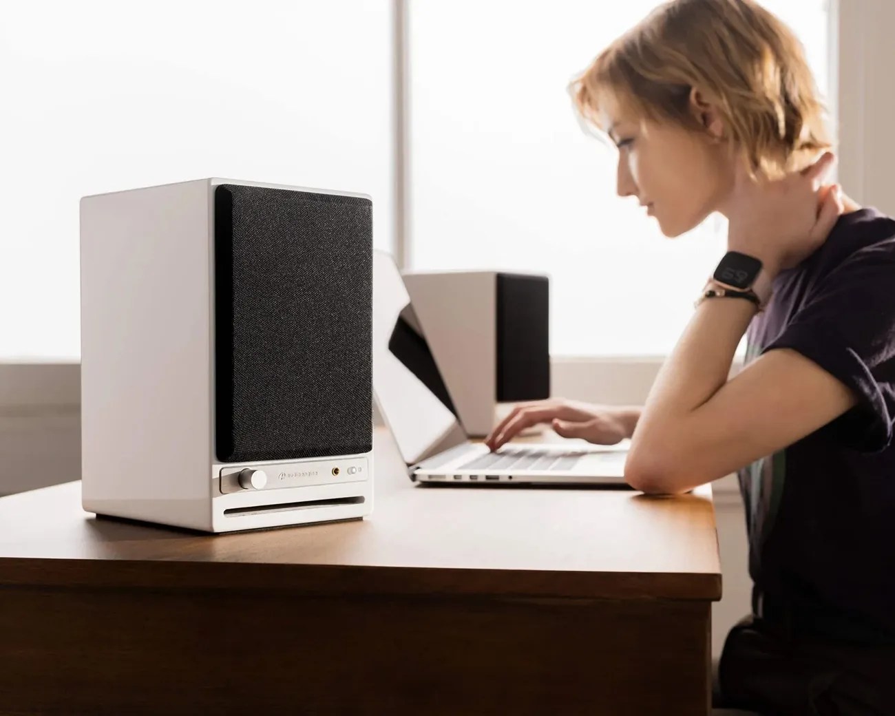 White desktop speaker with black fabric front and silver control knob on a wooden desk near a person using a laptop.