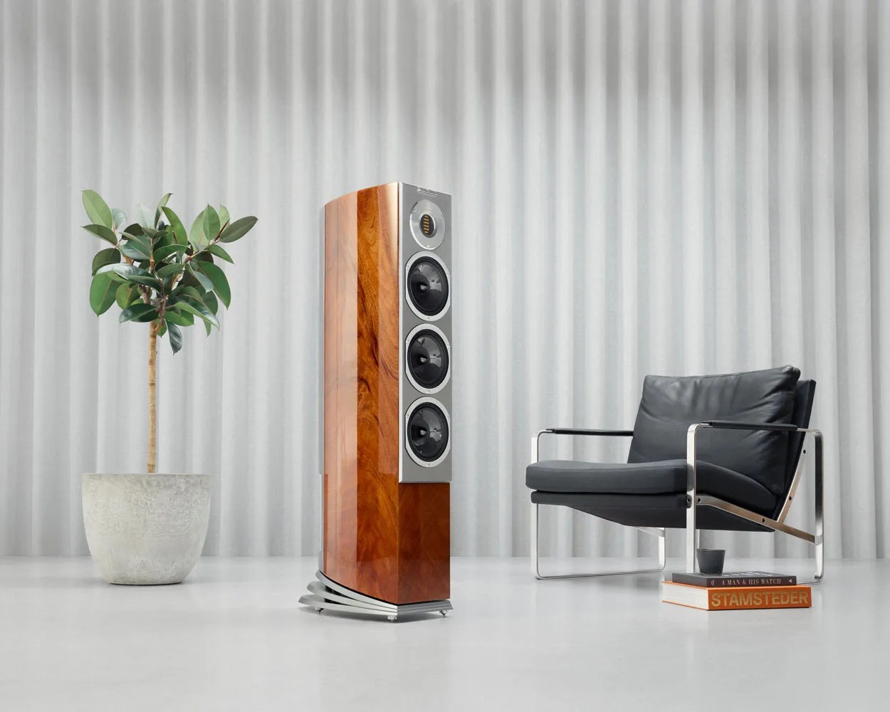 Tall wooden floor speaker with silver front panel and three black drivers next to a black leather chair and potted plant.