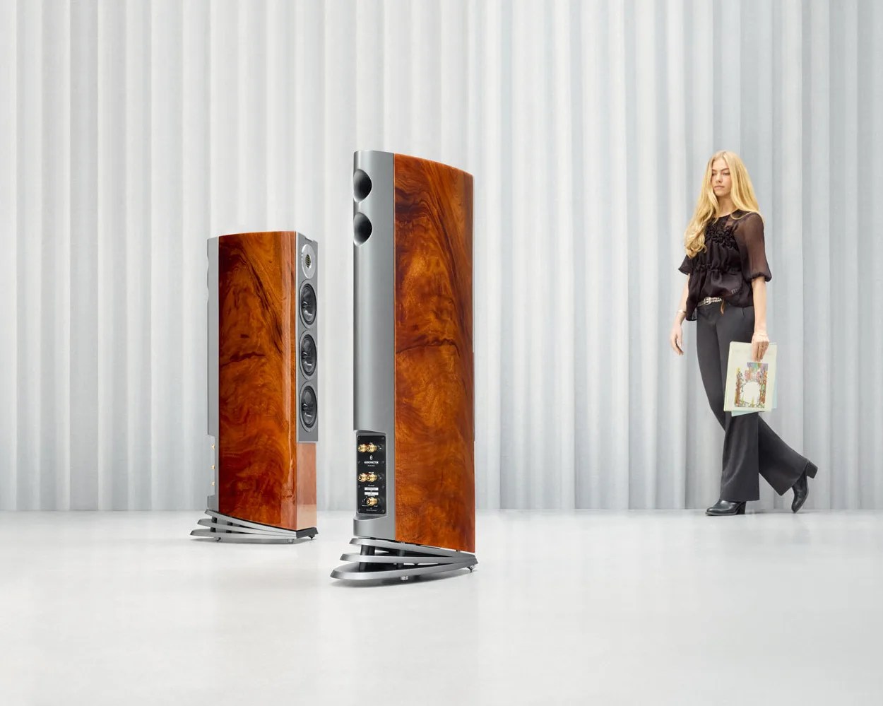 Two tall, polished wood and metal floor-standing speakers with curved bases in a white room, a person holding a vinyl record stands nearby.