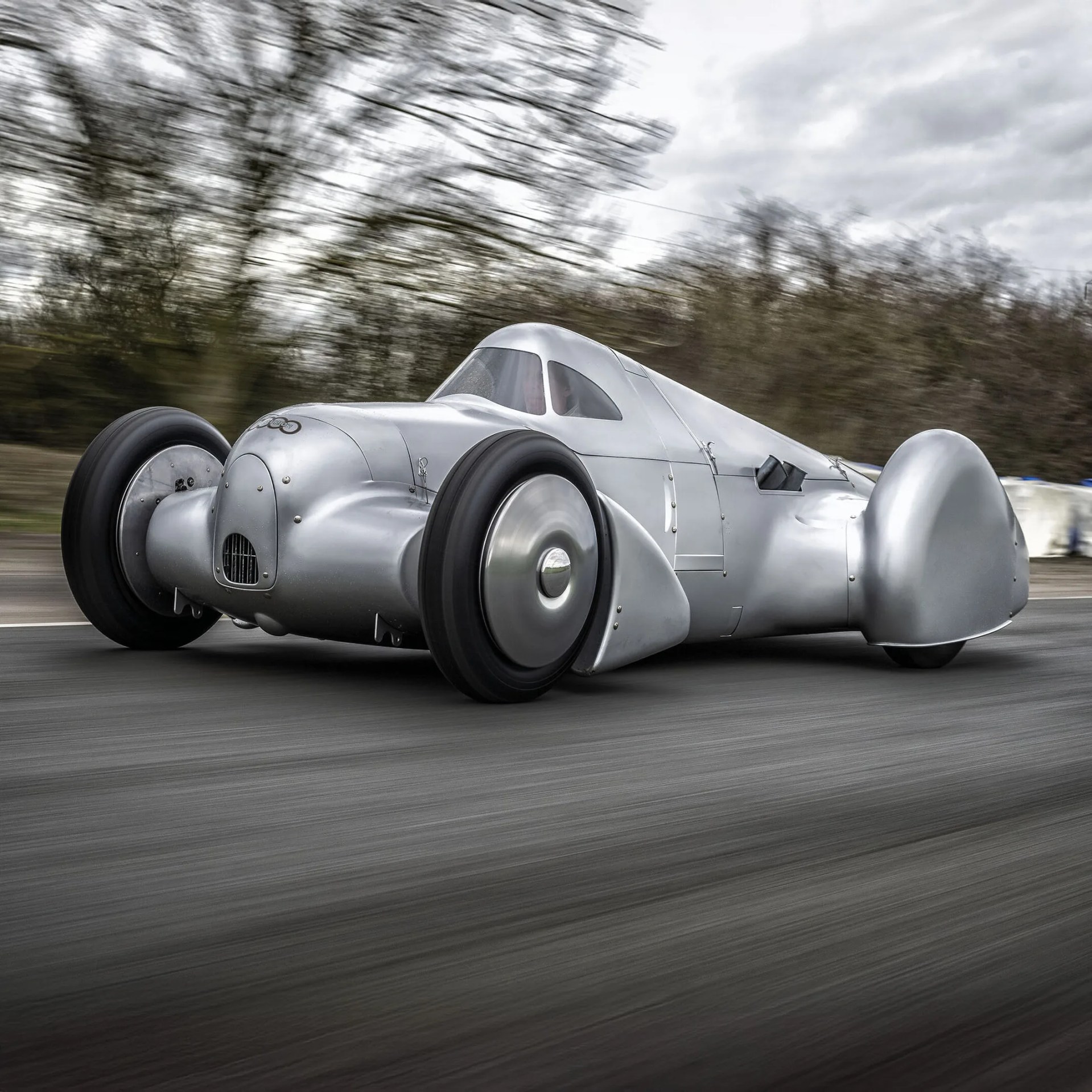 Streamlined silver vintage race car with covered wheels speeding on a road with blurred trees in the background.