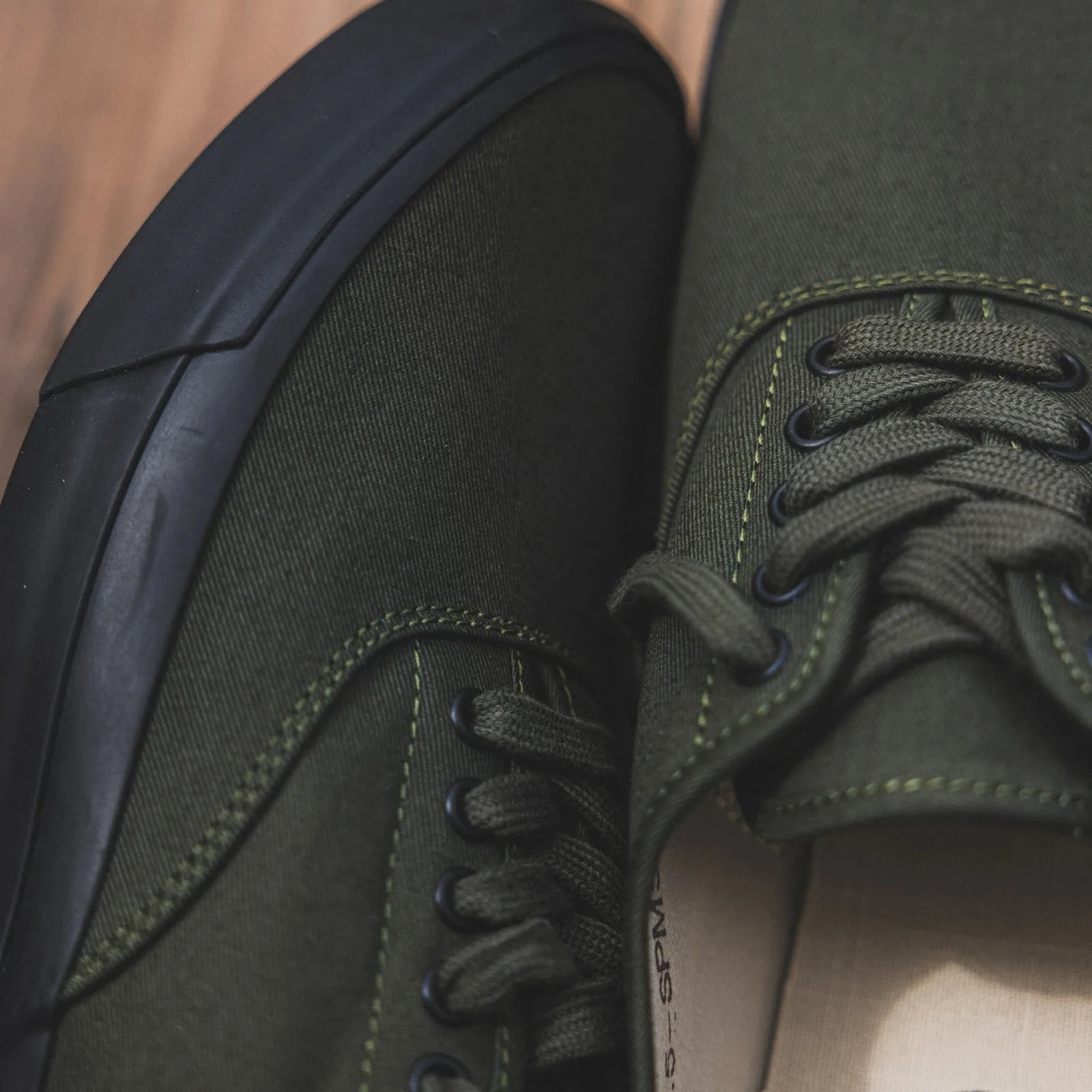 Close-up of olive green canvas sneakers with black rubber soles and matching laces on a wooden floor.