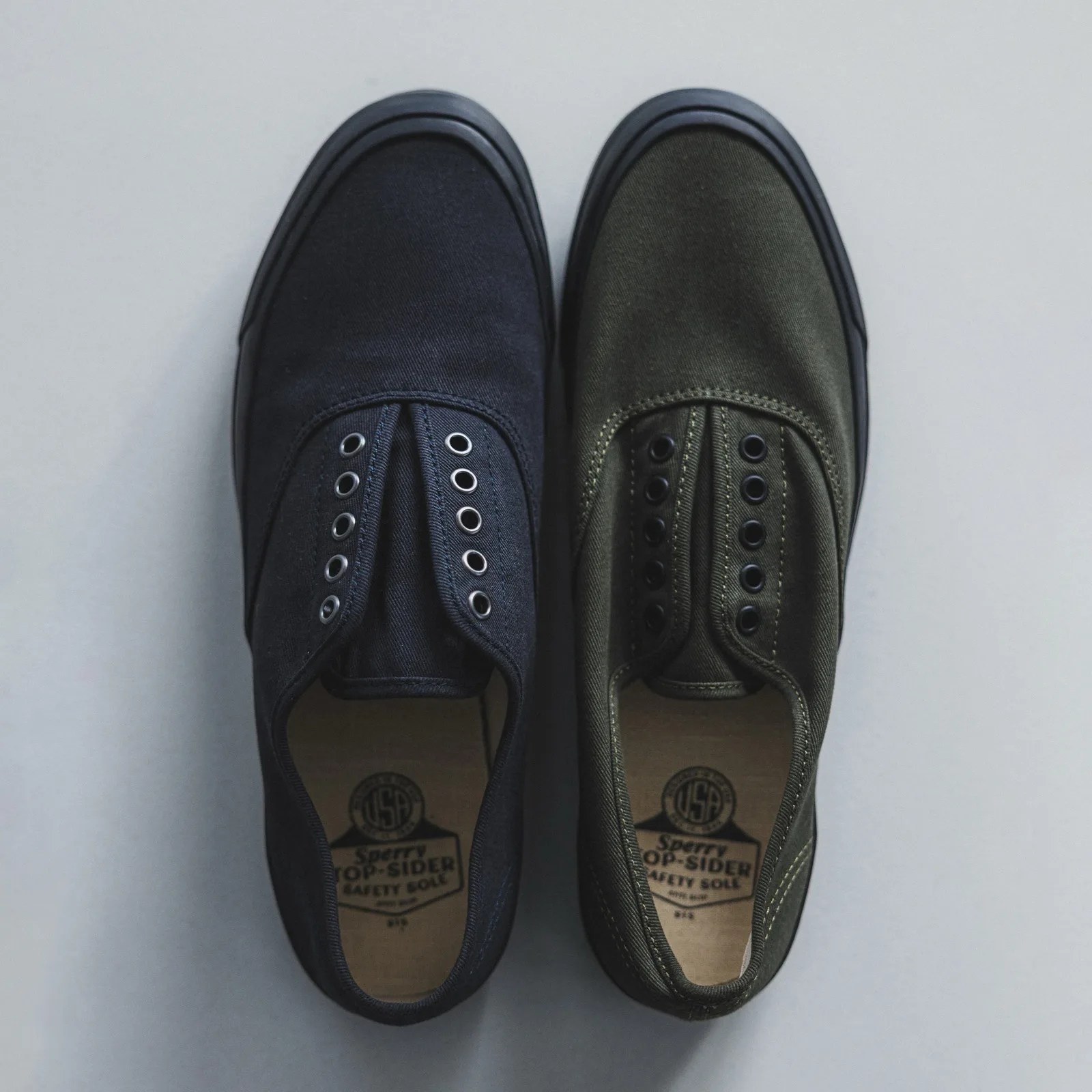 Pair of slip-on canvas shoes, one black and one olive green, with black rubber soles and metal eyelets.