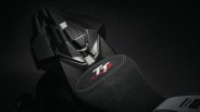 Black motorcycle seat with white and red "TT" logo and carbon fiber rear section under low light.