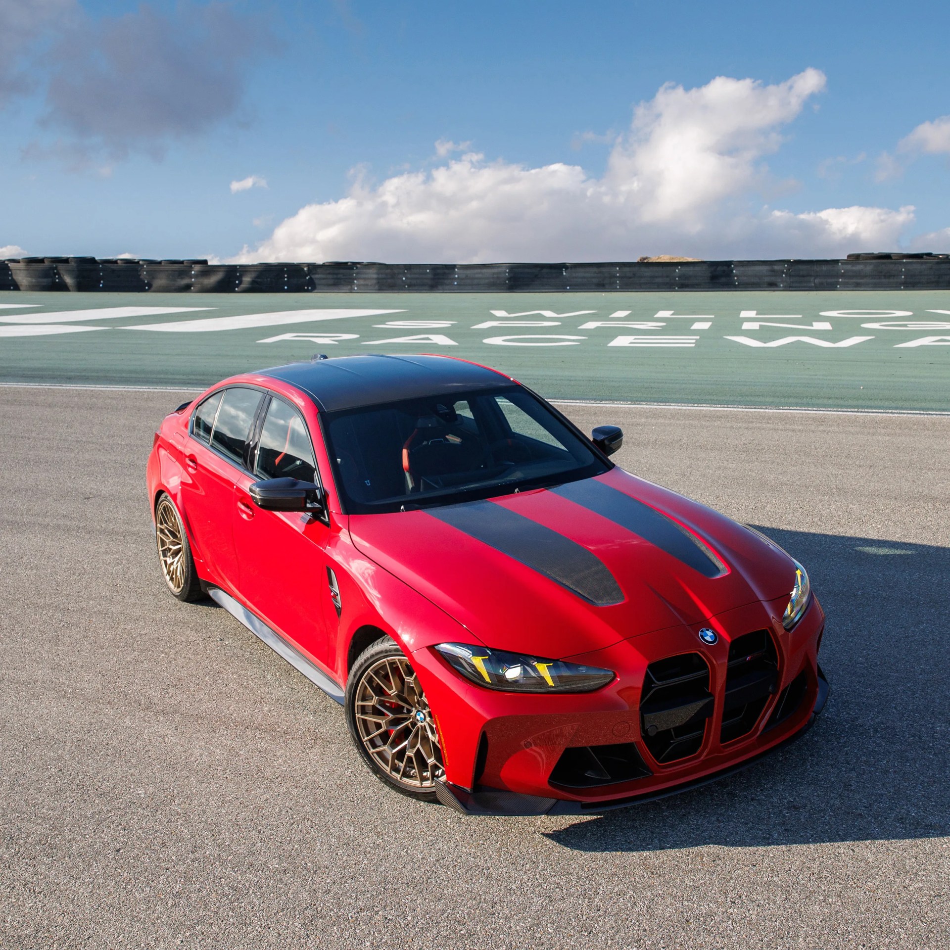 Red BMW sedan with black racing stripes and bronze wheels parked on a raceway track.