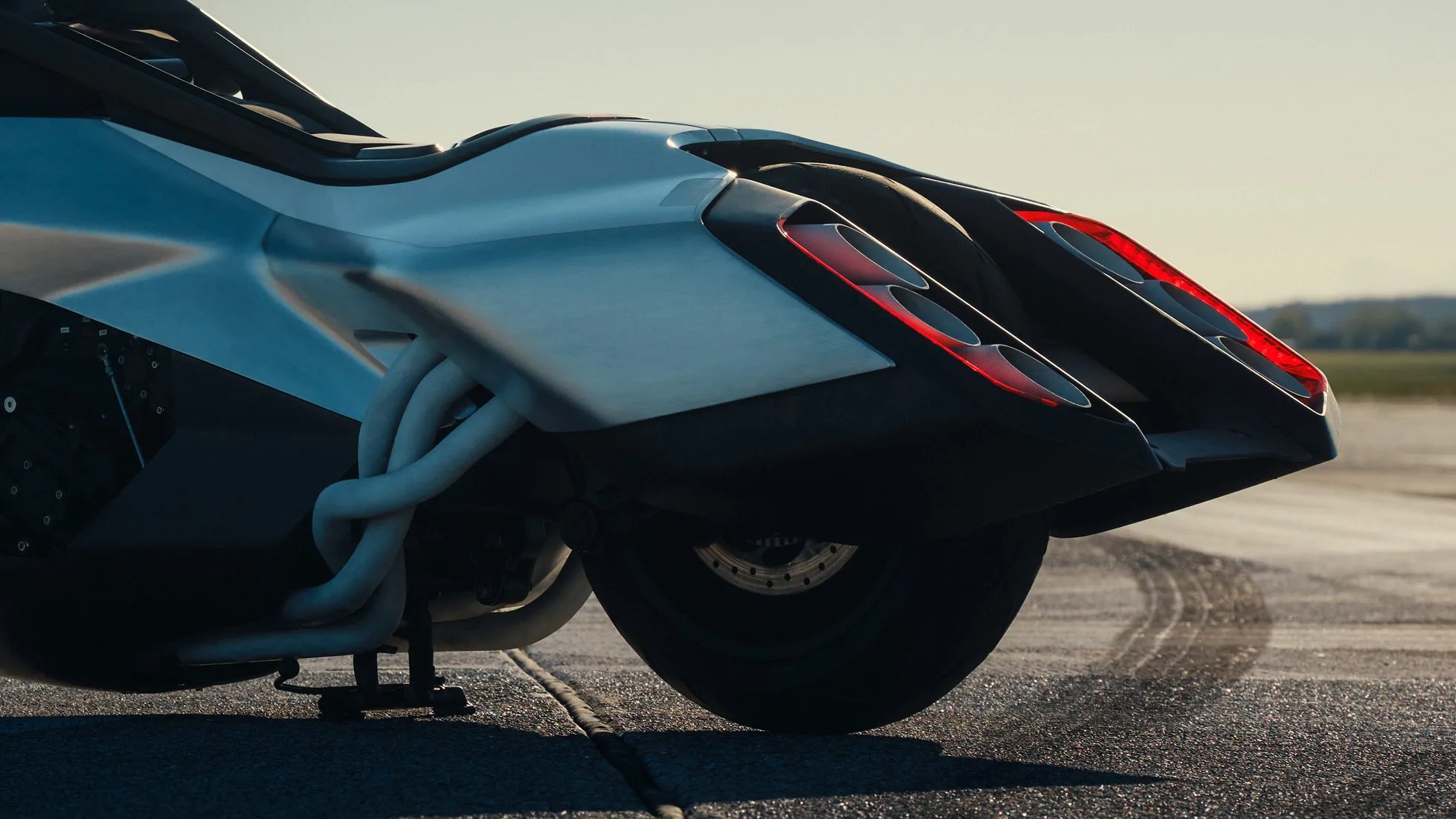 Rear view of a futuristic motorcycle with dual exhaust pipes and red-accented tail lights on a paved surface.