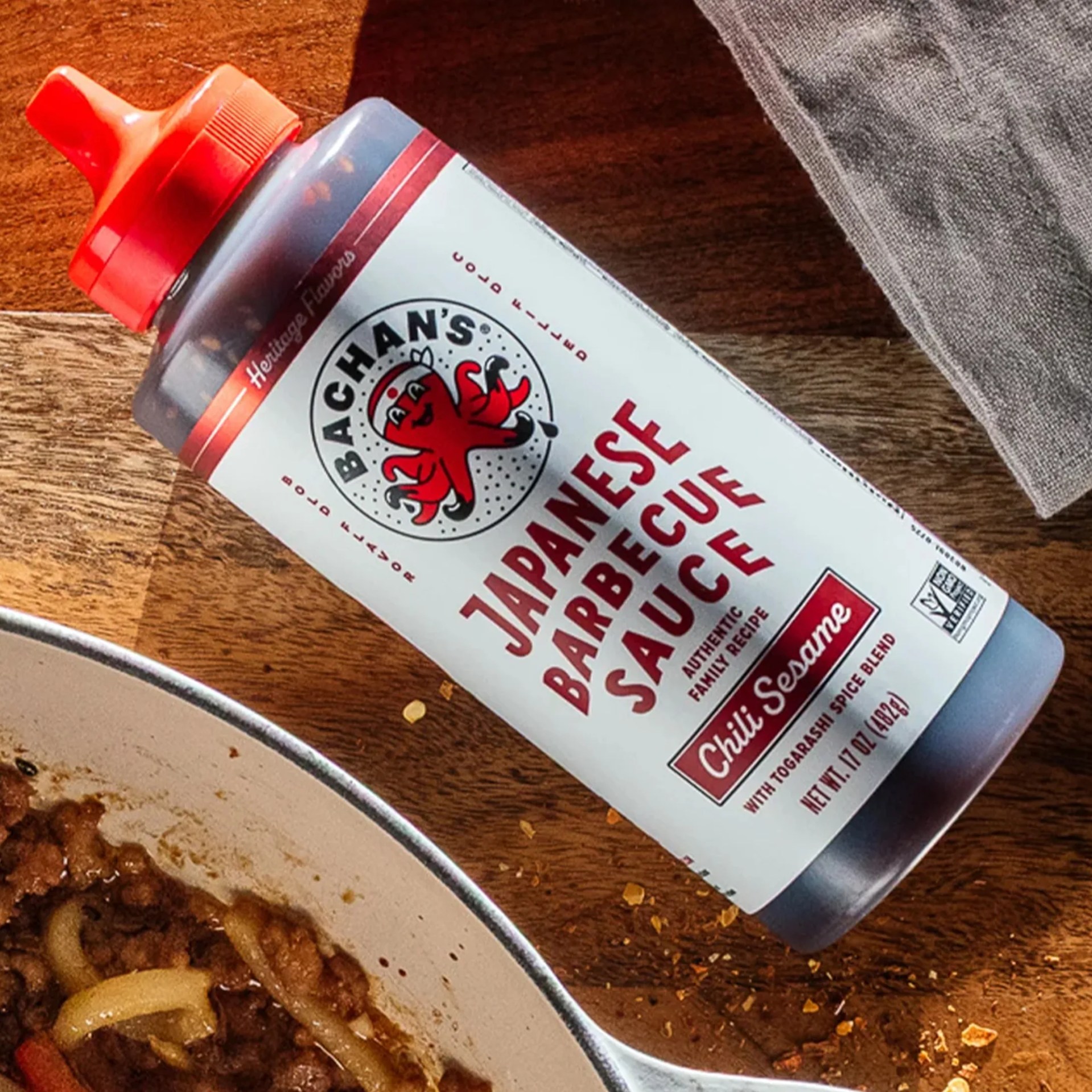 Bottle of Bachan's Japanese Barbecue Sauce with chili sesame flavor on a wooden surface next to a bowl of cooked food.