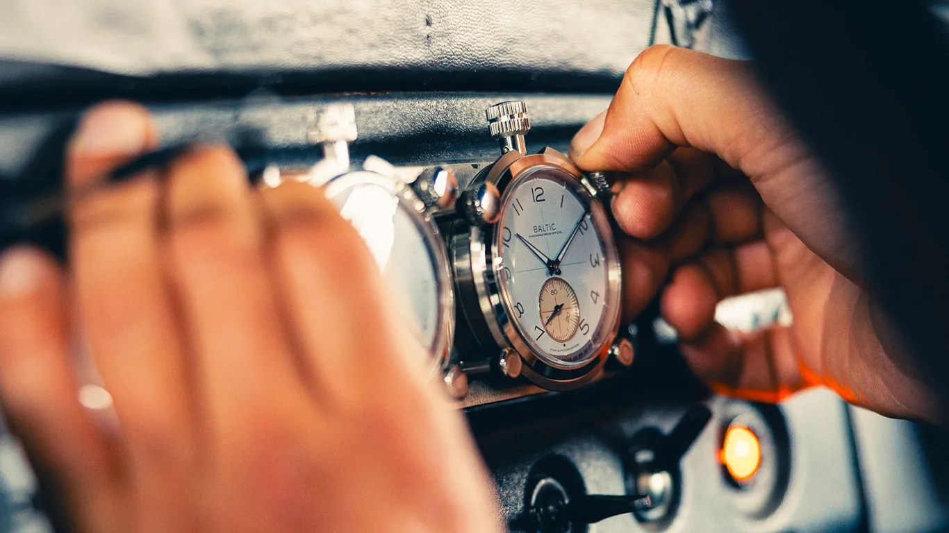 Hands adjusting a silver Baltic stopwatch mounted on a black dashboard.
