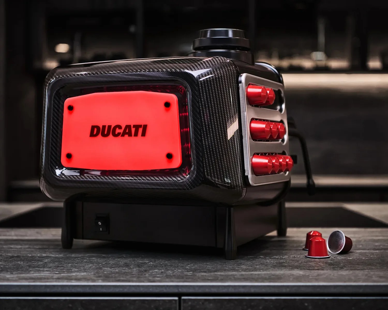 Black carbon fiber espresso machine with red Ducati logo and red coffee capsules on a countertop.