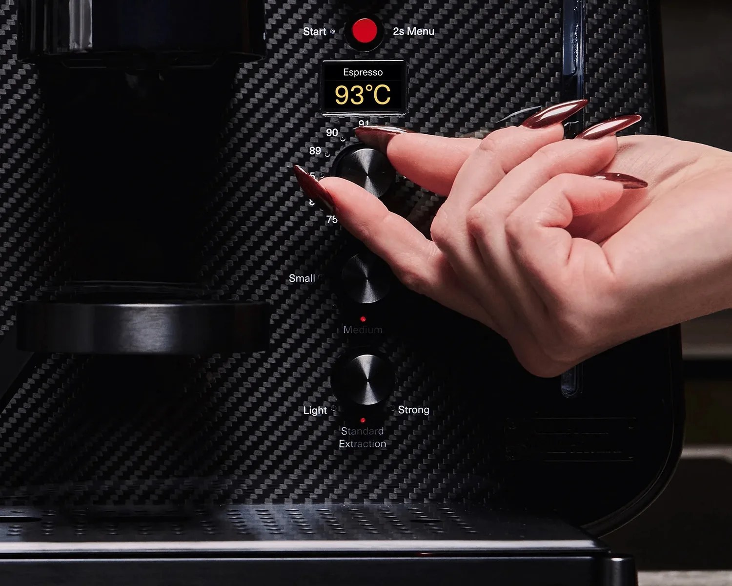 Hand with long dark red nails adjusting the temperature dial on a black carbon fiber espresso machine showing 93°C.