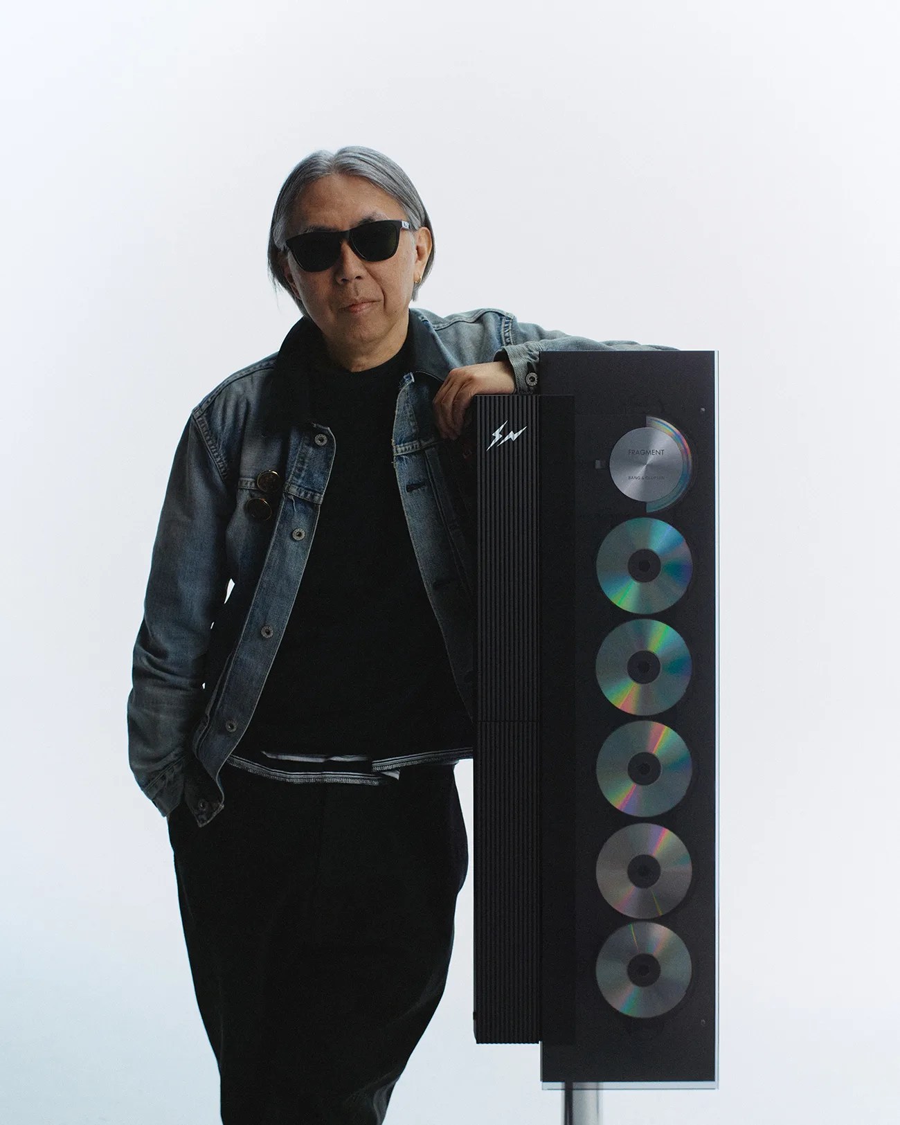 Person wearing sunglasses and a denim jacket leaning on a tall black speaker with multiple CDs displayed vertically.