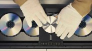Hands wearing white gloves placing a silver disc labeled "BANG & OLUFSEN" into a black multi-disc player with other discs.