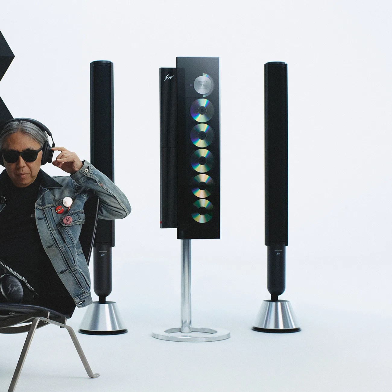 Black tall speakers with silver bases and a central speaker stand featuring six CDs mounted vertically, next to a person wearing headphones and a denim jacket.