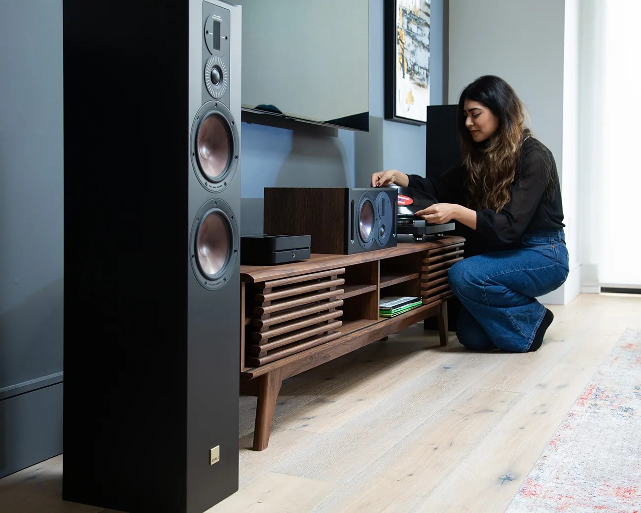 Tall black floor-standing speaker with copper-colored drivers next to a wooden media console with a woman placing a vinyl record on a turntable.