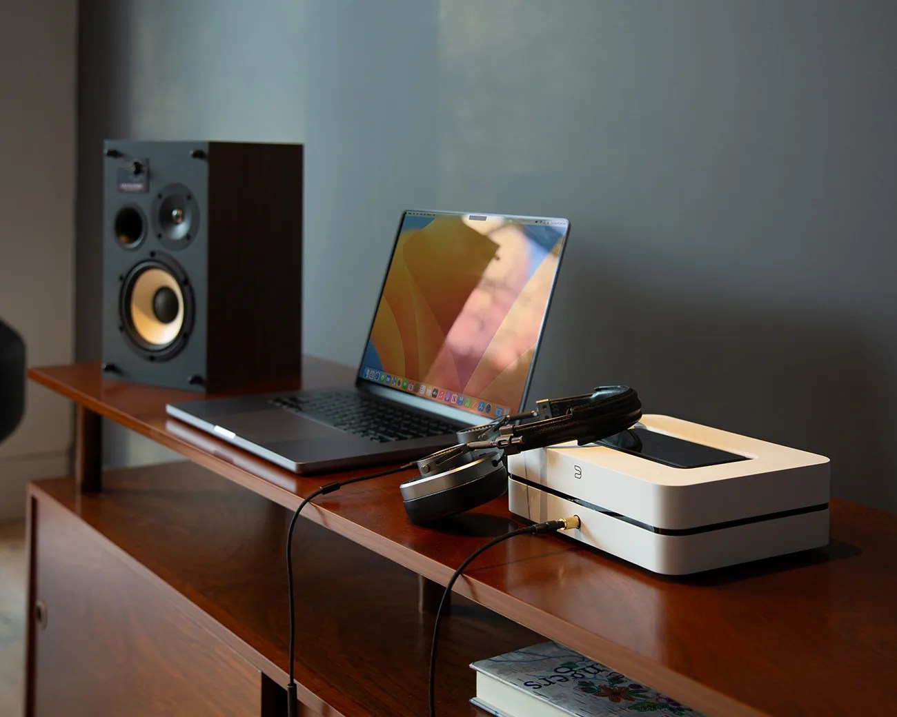 Wooden speaker, open laptop, white audio device, and black wired headphones on a wooden shelf.