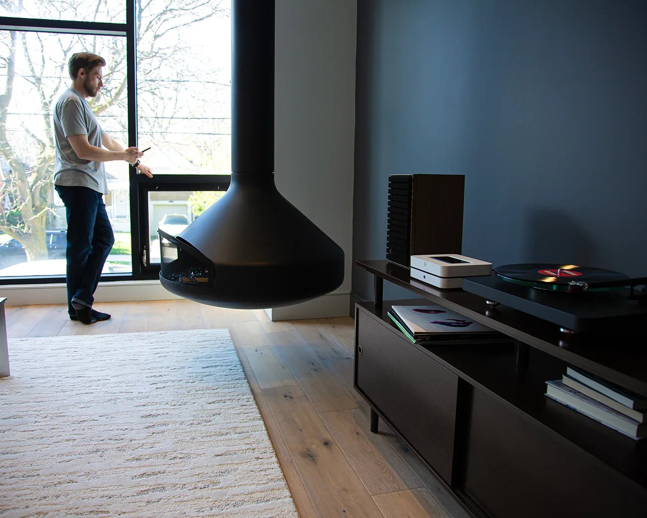 Man in a gray shirt and dark pants standing near a large window beside a hanging black fireplace in a modern living room.