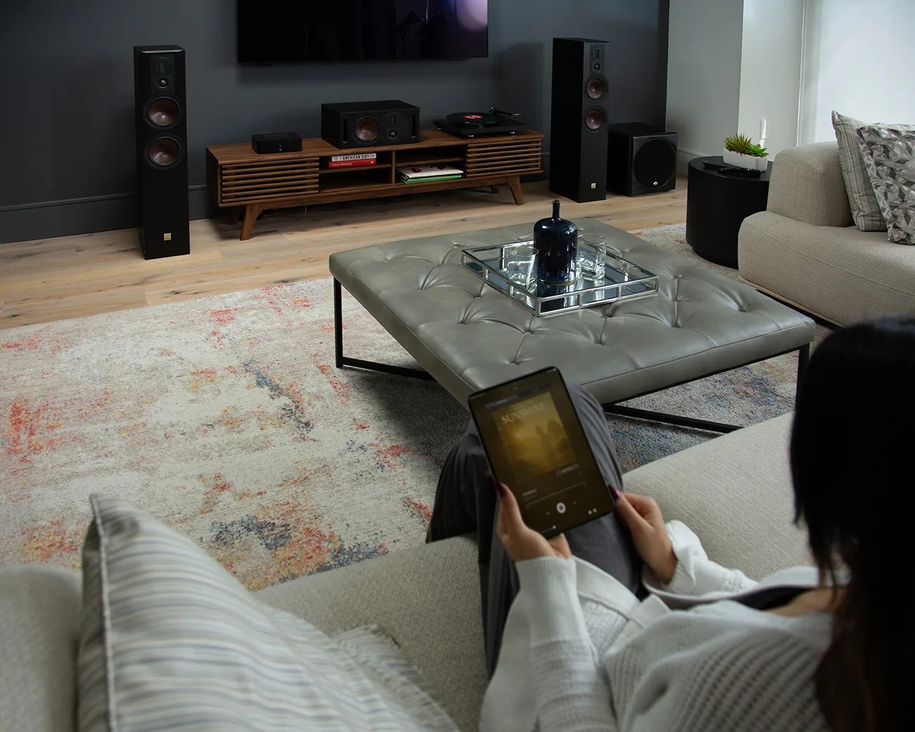 Person holding a tablet while sitting on a beige sofa in a modern living room with a tufted gray ottoman and wooden media console.