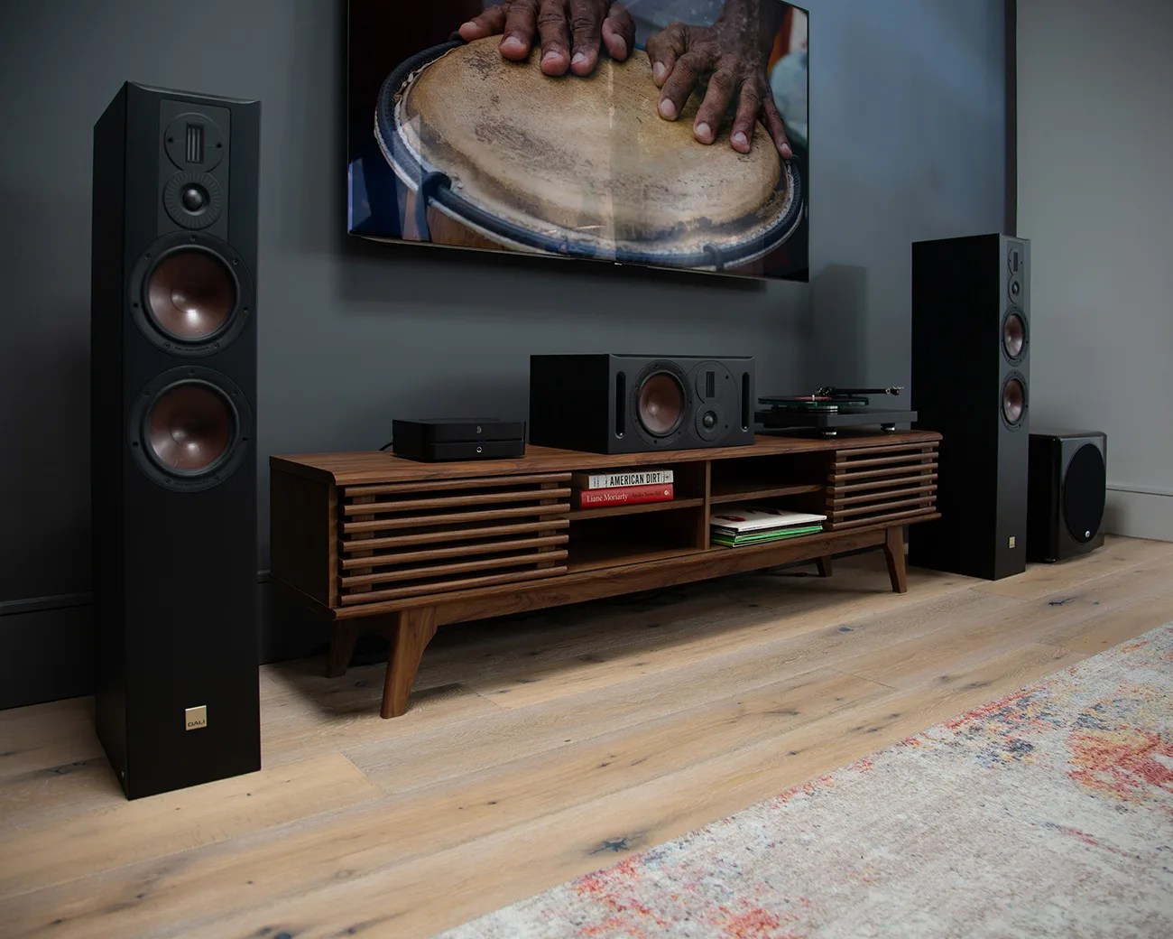 Home entertainment setup with two tall black floor-standing speakers, a wooden media console holding a center speaker, a turntable, and a subwoofer on light wood flooring.