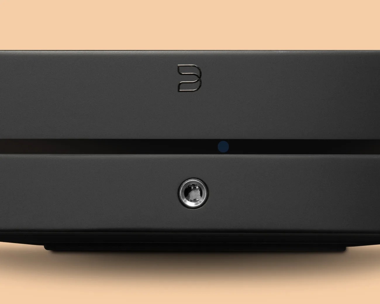 Front view of a black electronic device with a slot and a circular port below it on a beige background.