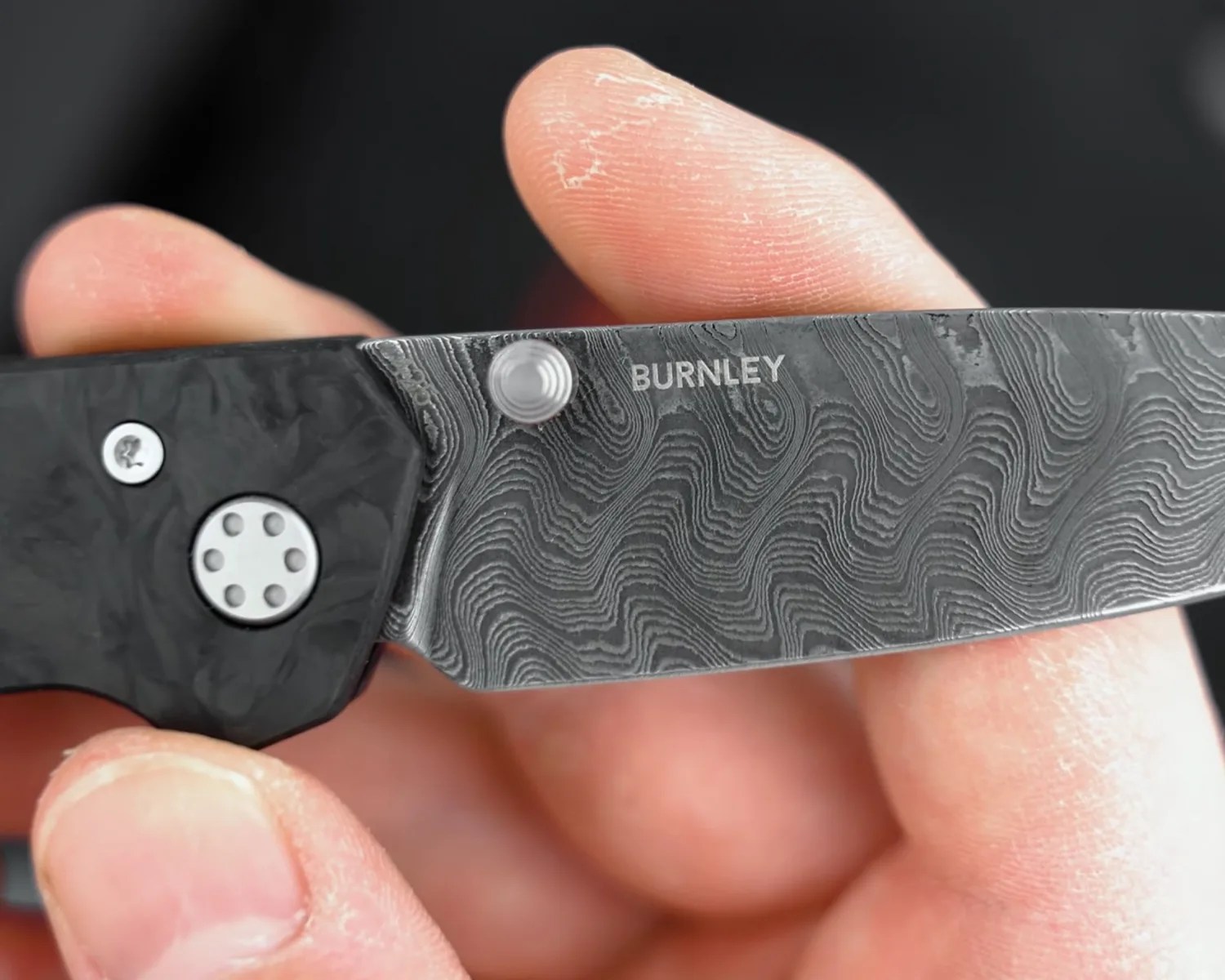 Close-up of a Damascus steel knife blade with "BURNLEY" engraved, held between fingers.