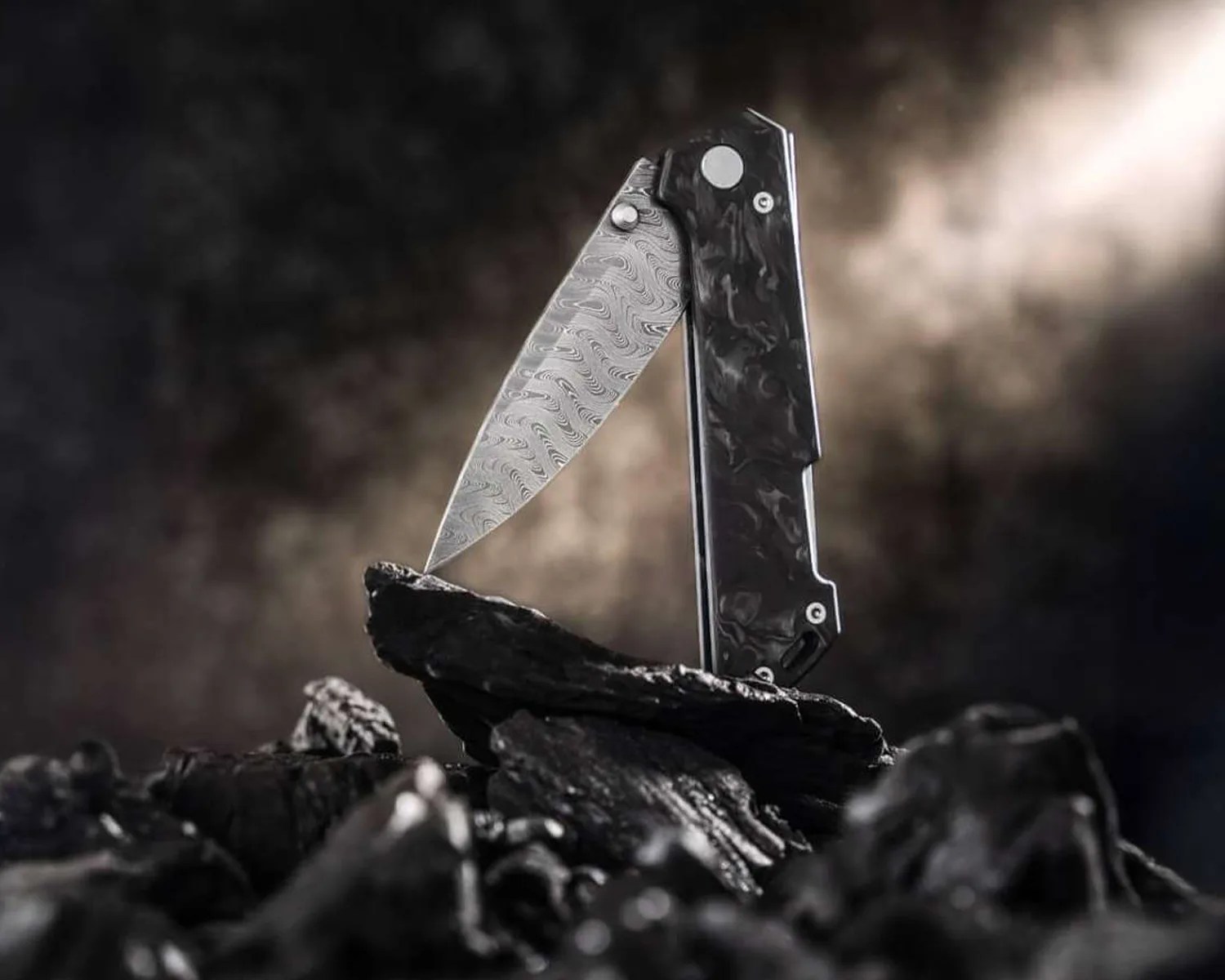 Folding knife with a patterned blade and black marbled handle standing open on dark rocks.