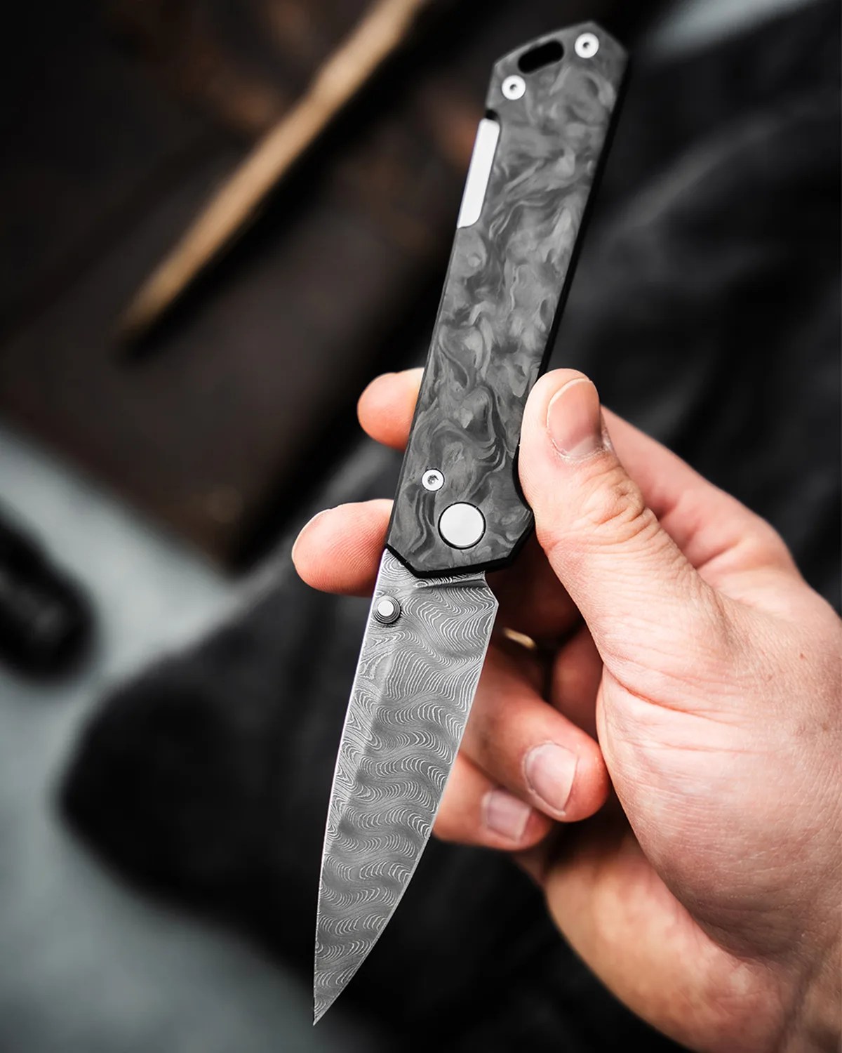 Hand holding a folding knife with a patterned Damascus steel blade and a black marbled handle.