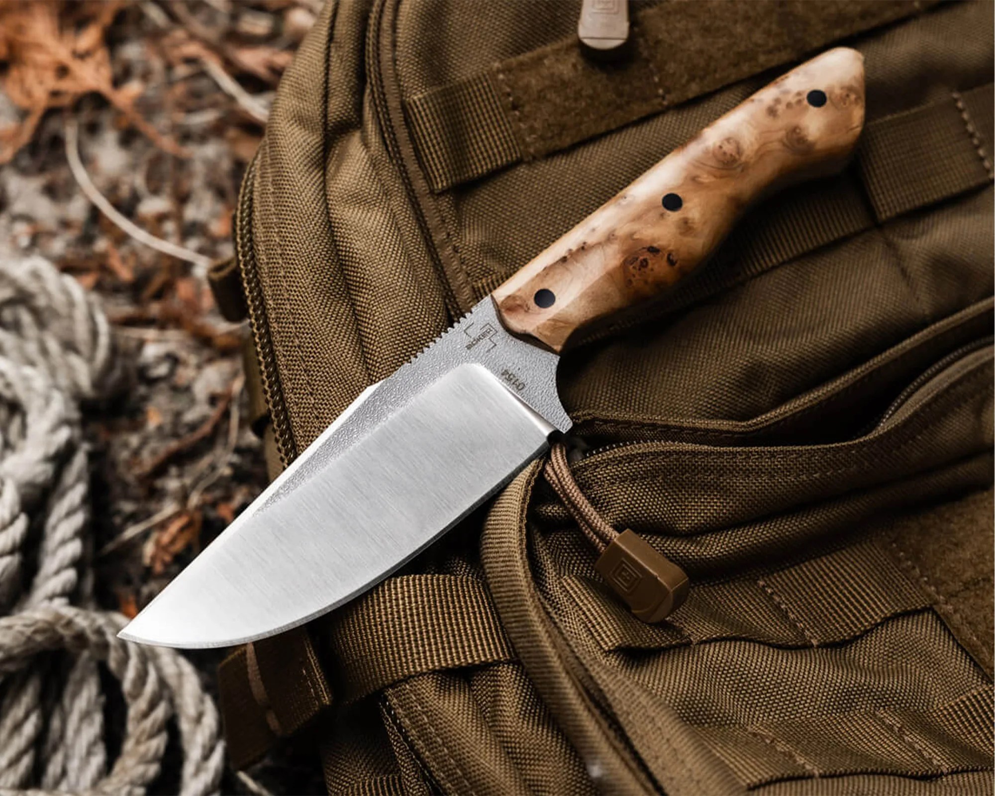 Knife with a polished steel blade and a light wood handle resting on a brown fabric backpack.