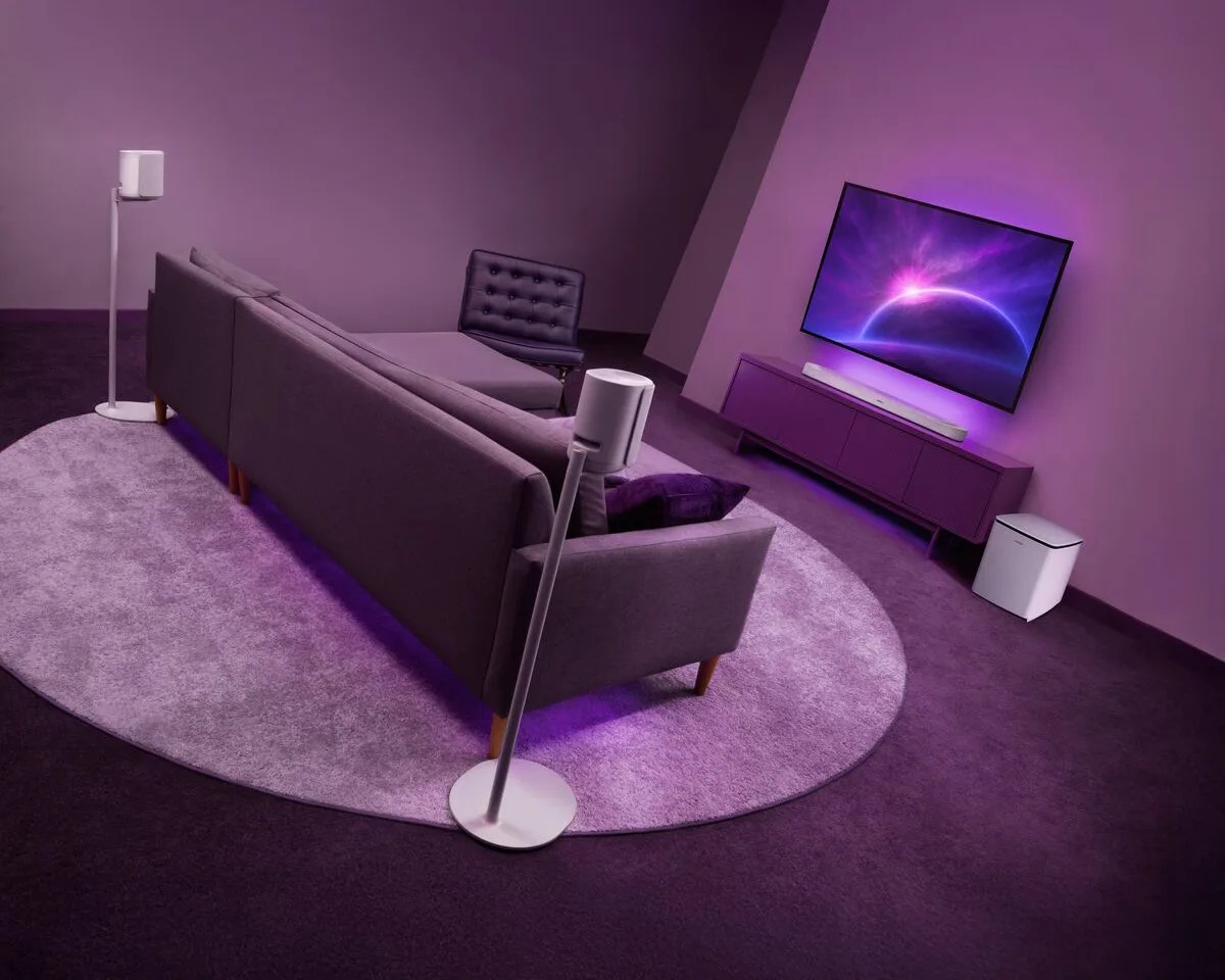 Modern living room with a gray sectional sofa, black chair, white floor speakers, purple round rug, wall-mounted TV, and white subwoofer.