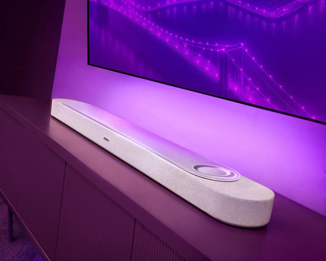 White Bose soundbar with fabric cover and circular control panel on a purple cabinet under a wall-mounted TV.