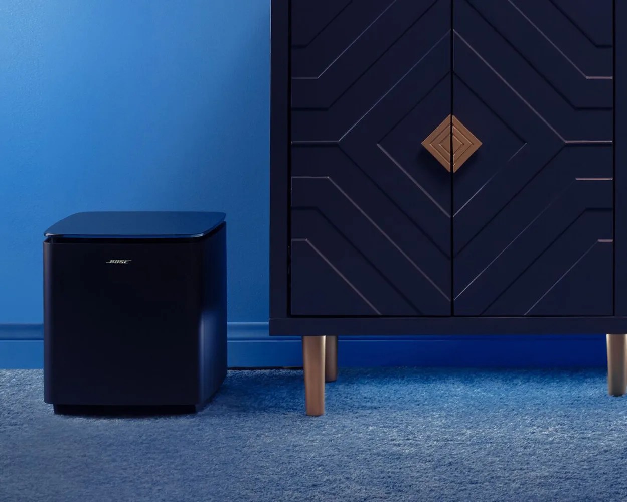 Black Bose subwoofer speaker on a blue carpet next to a dark blue cabinet with gold legs and a geometric gold handle.