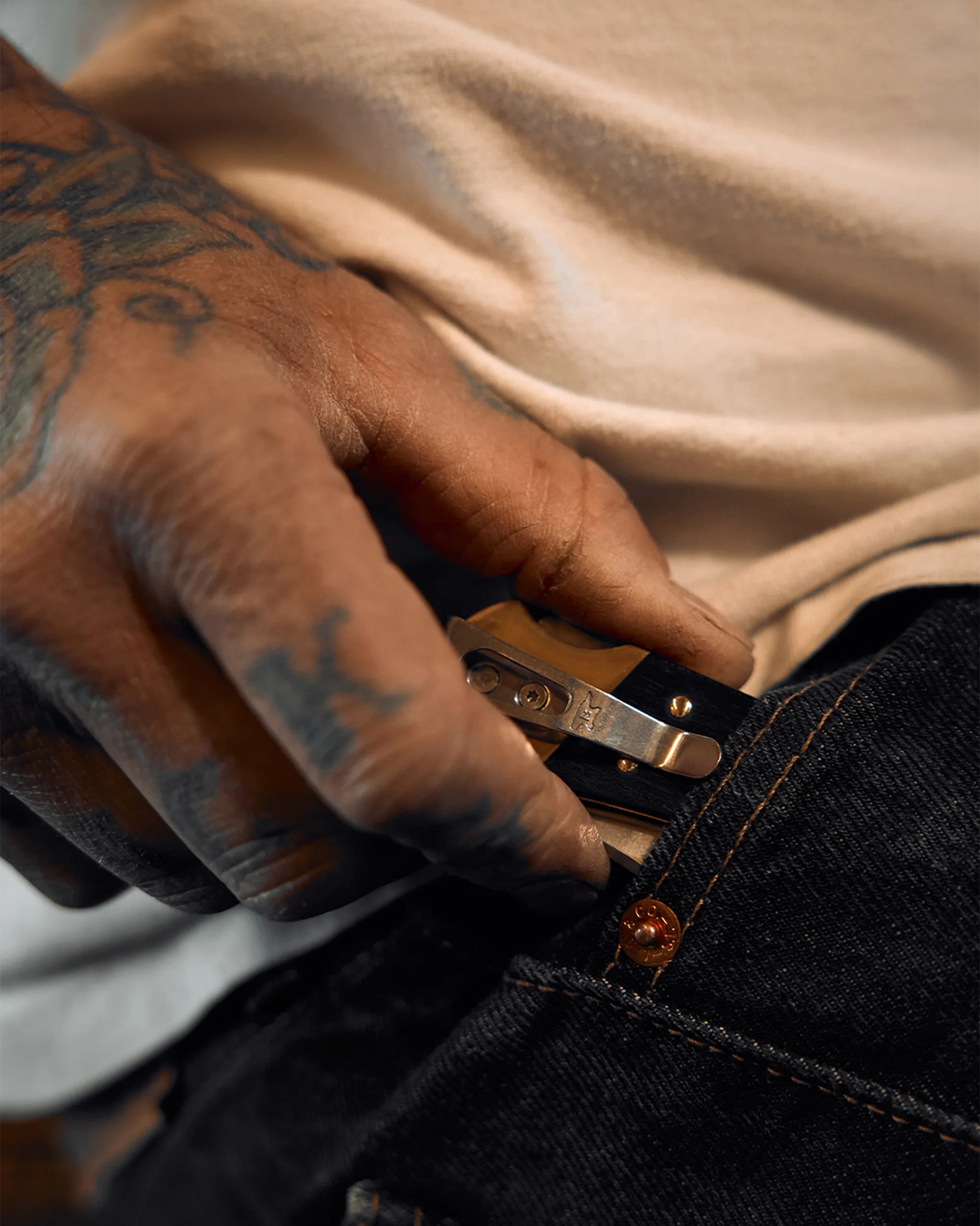 Tattooed hand holding a black and brown folding knife clipped to dark denim jeans pocket.