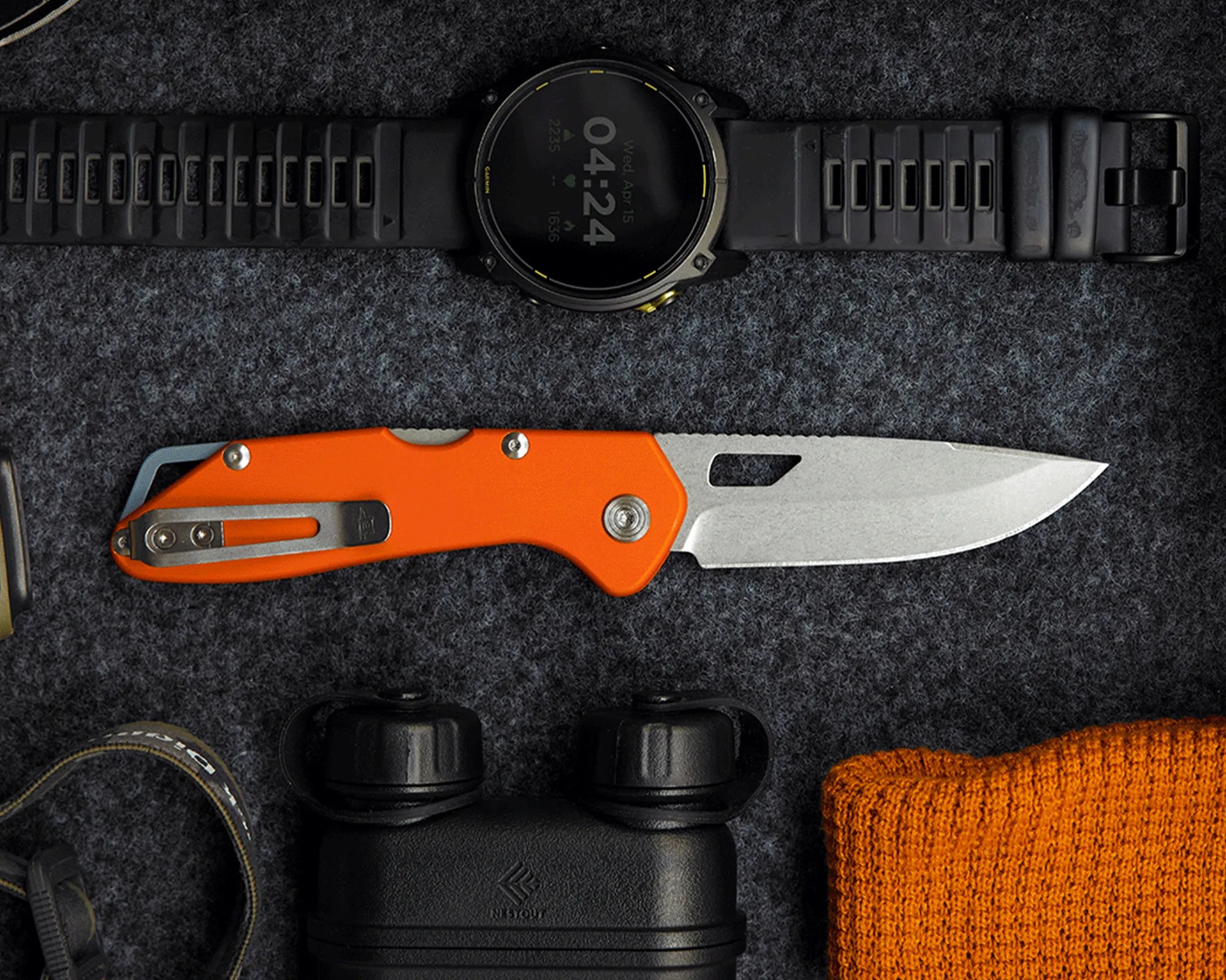Black digital wristwatch with a black rubber strap and an orange-handled folding knife with a silver blade on a dark fabric surface.