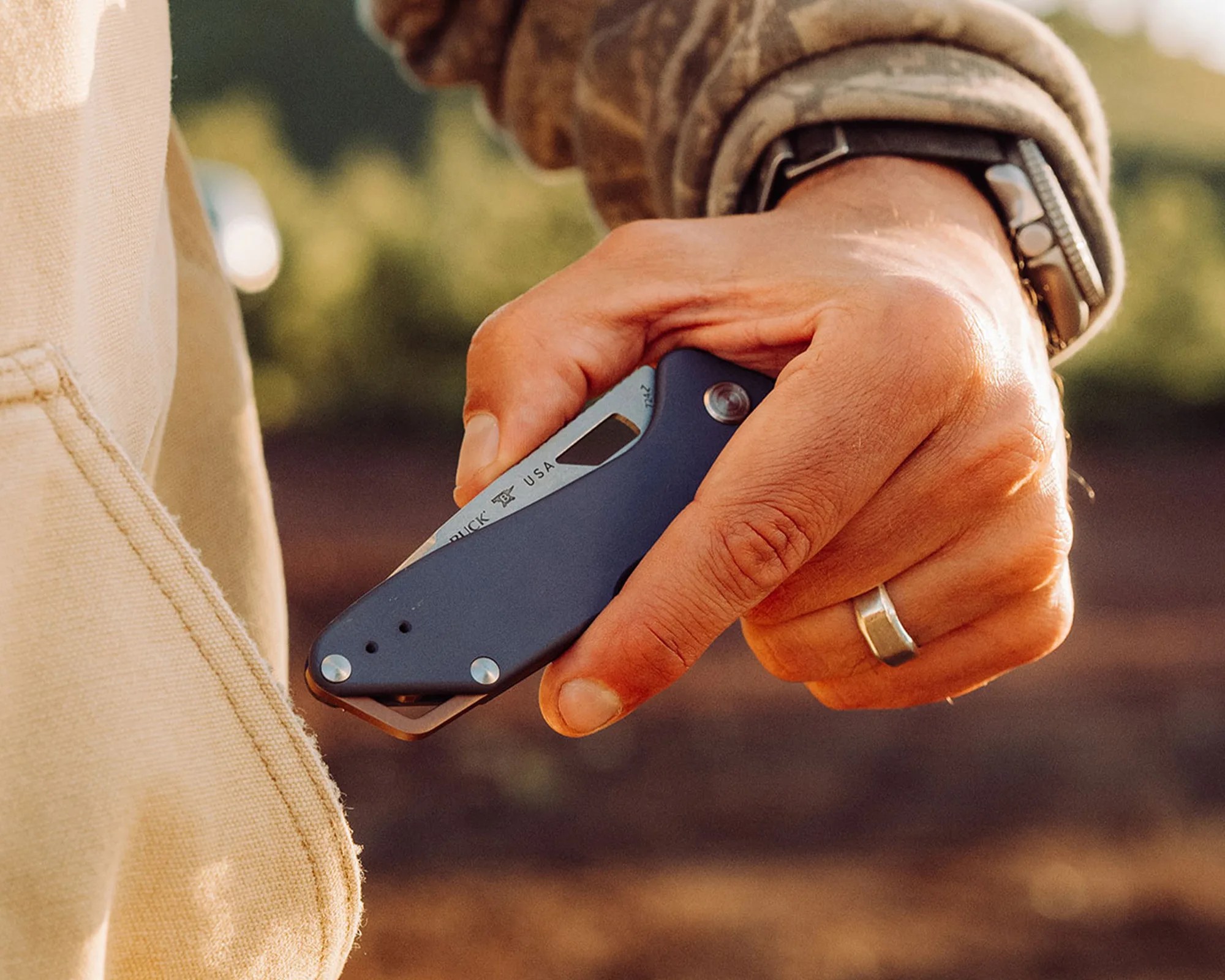 Hand holding a closed dark blue folding knife with visible "BUCK USA" engraving near beige fabric.