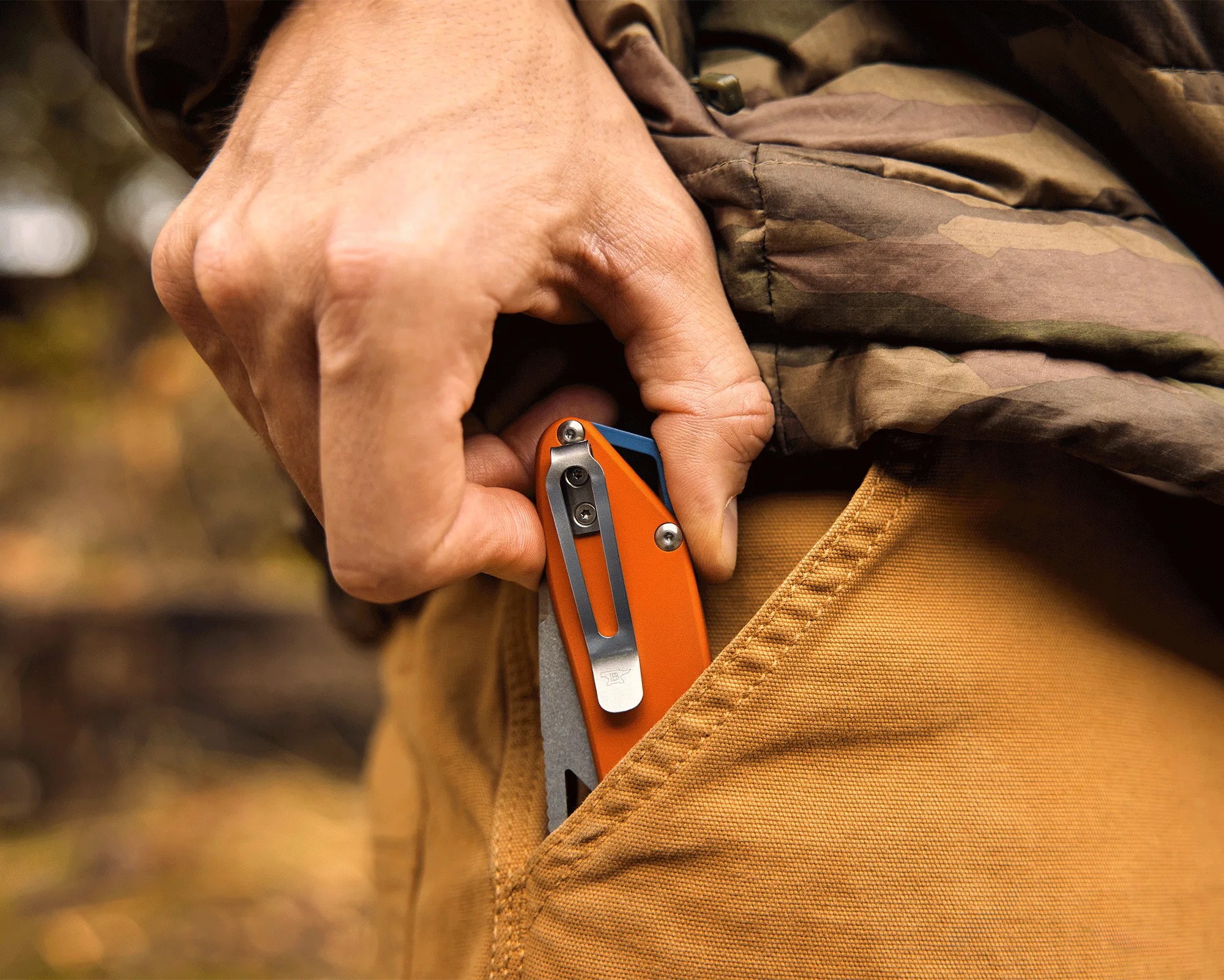 Hand holding an orange folding knife clipped to the pocket of mustard-colored pants and camo jacket.