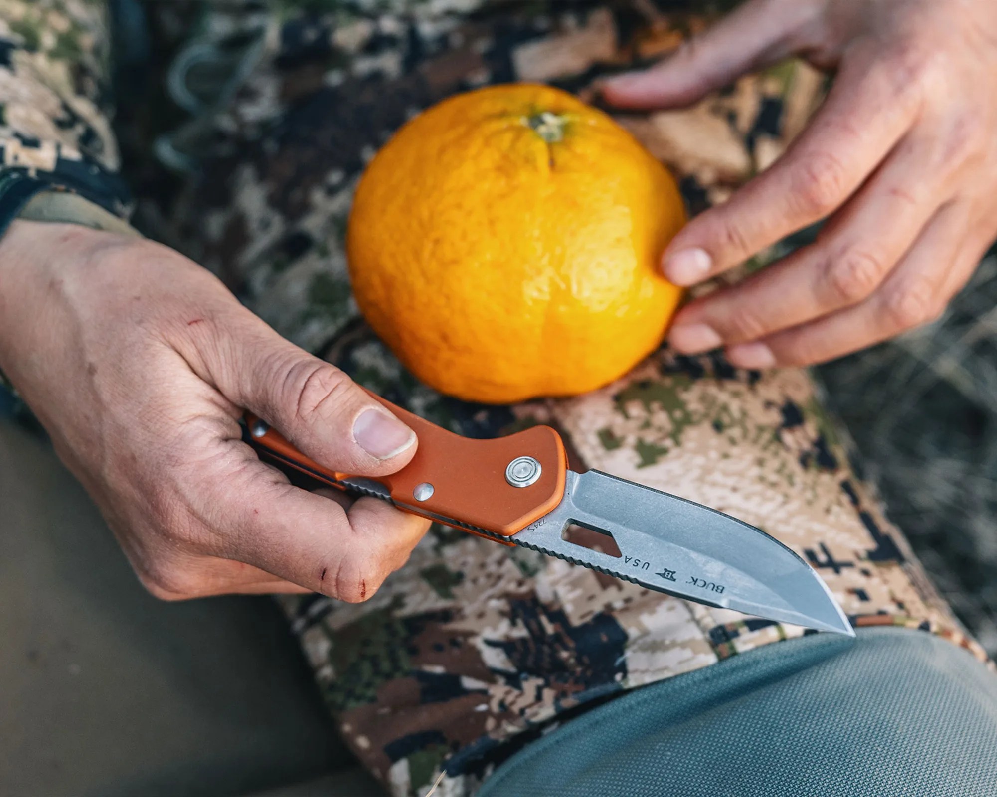 Hands holding an orange-handled Buck USA folding knife with an orange on camouflage fabric.