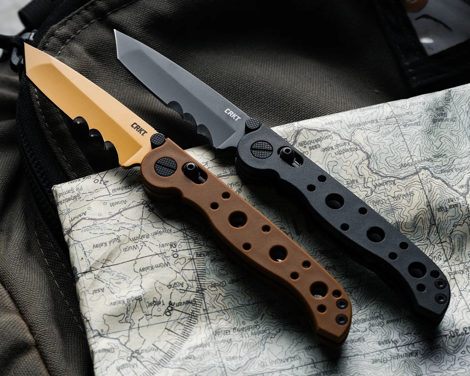 Two CRKT folding knives with tanto blades, one with a tan blade and brown handle, the other with a black blade and black handle, on a map.