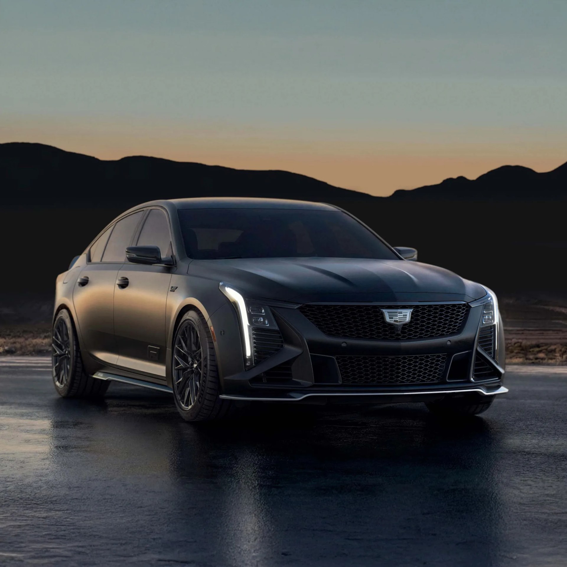 Matte black Cadillac sedan with vertical LED headlights parked on wet pavement at dusk.