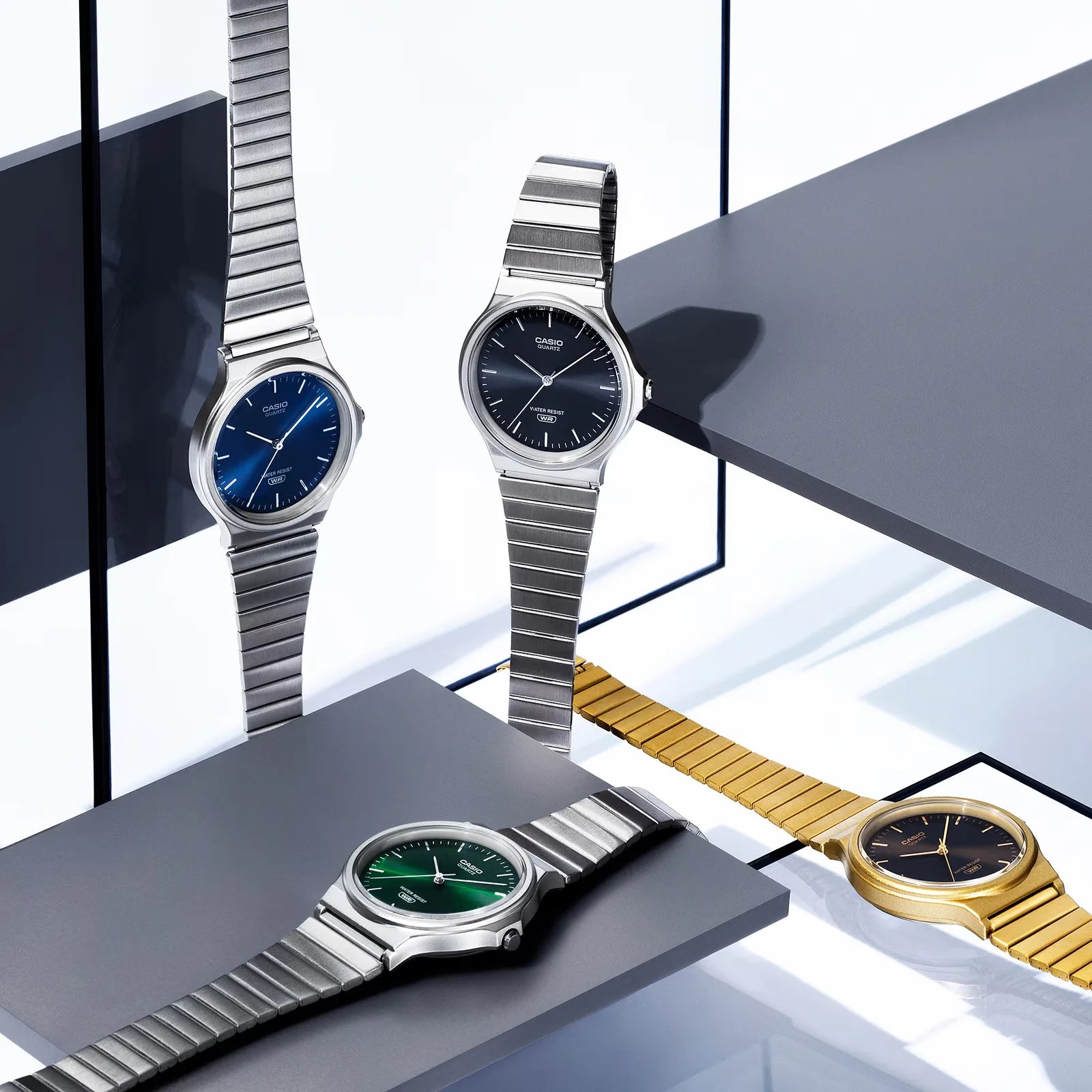 Four Casio quartz watches with metal bands in silver and gold, featuring blue, green, and black dials on gray platforms.