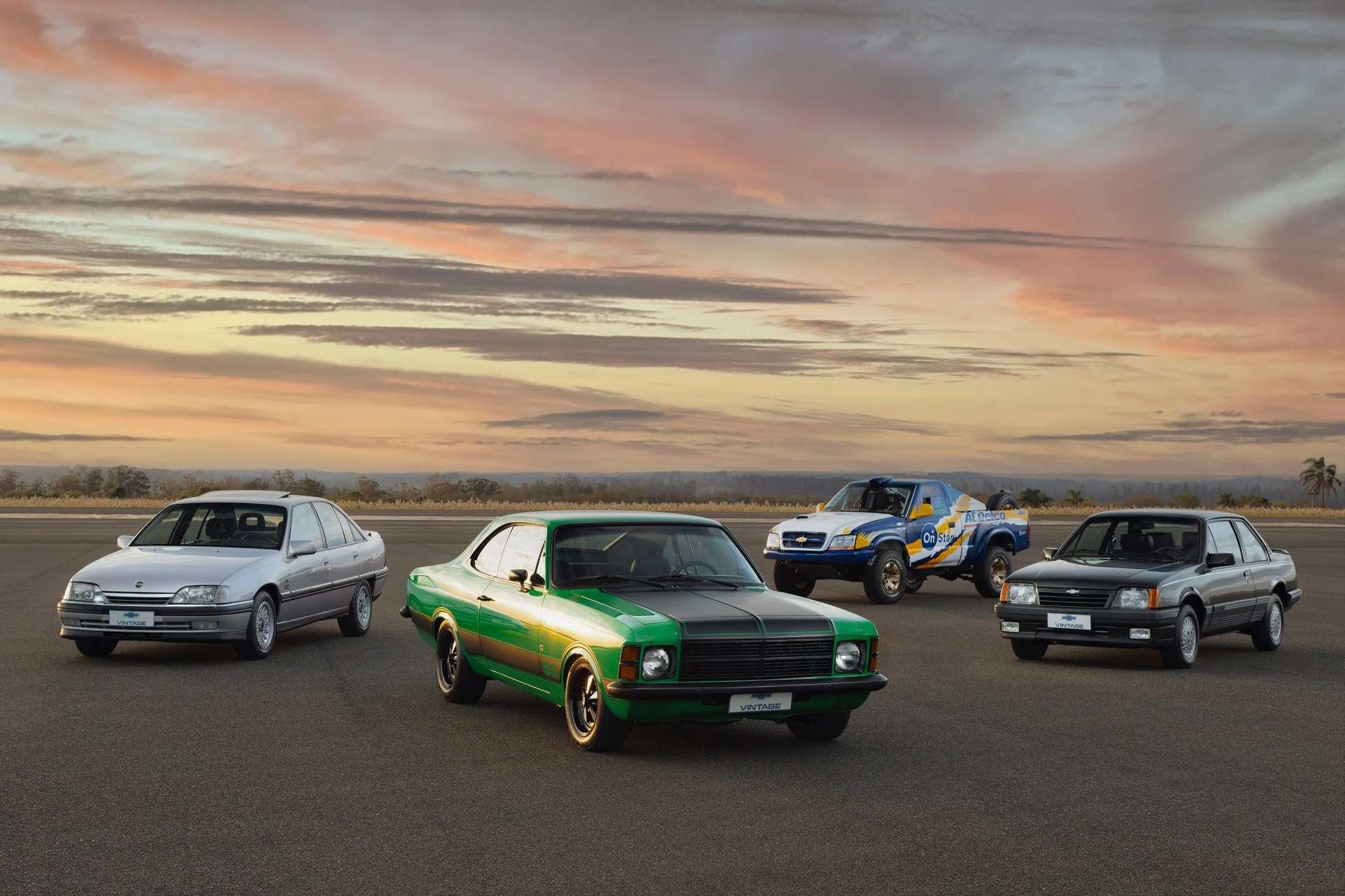 Four vintage cars parked on an open tarmac under a colorful sunset sky.