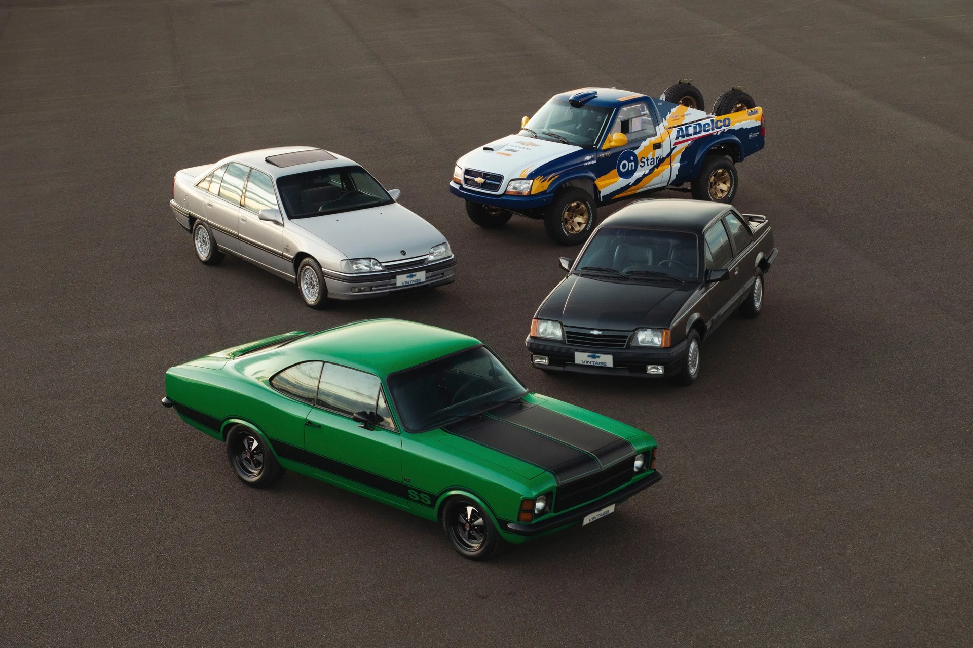 Four vintage cars including a green muscle car with black stripes, a silver sedan, a black compact car, and a blue and white pickup truck with yellow accents on an asphalt surface.