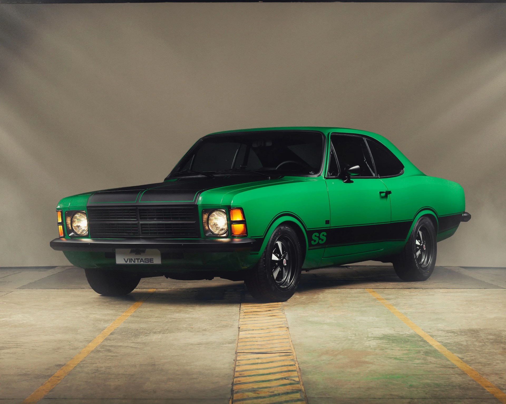 Green vintage muscle car with black racing stripes and SS logo on the side, parked indoors.