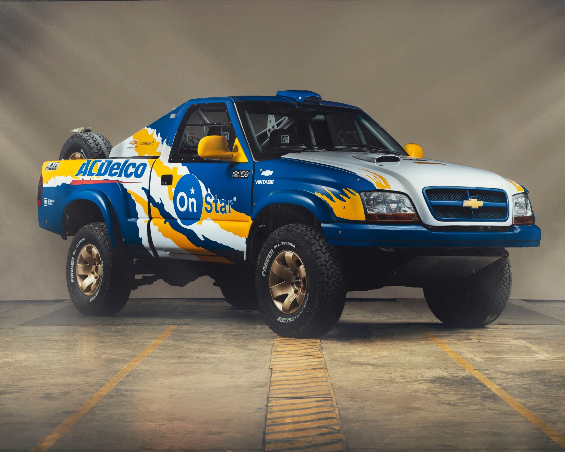 Blue, white, and yellow Chevrolet S10 off-road truck with sponsor decals and large all-terrain tires.