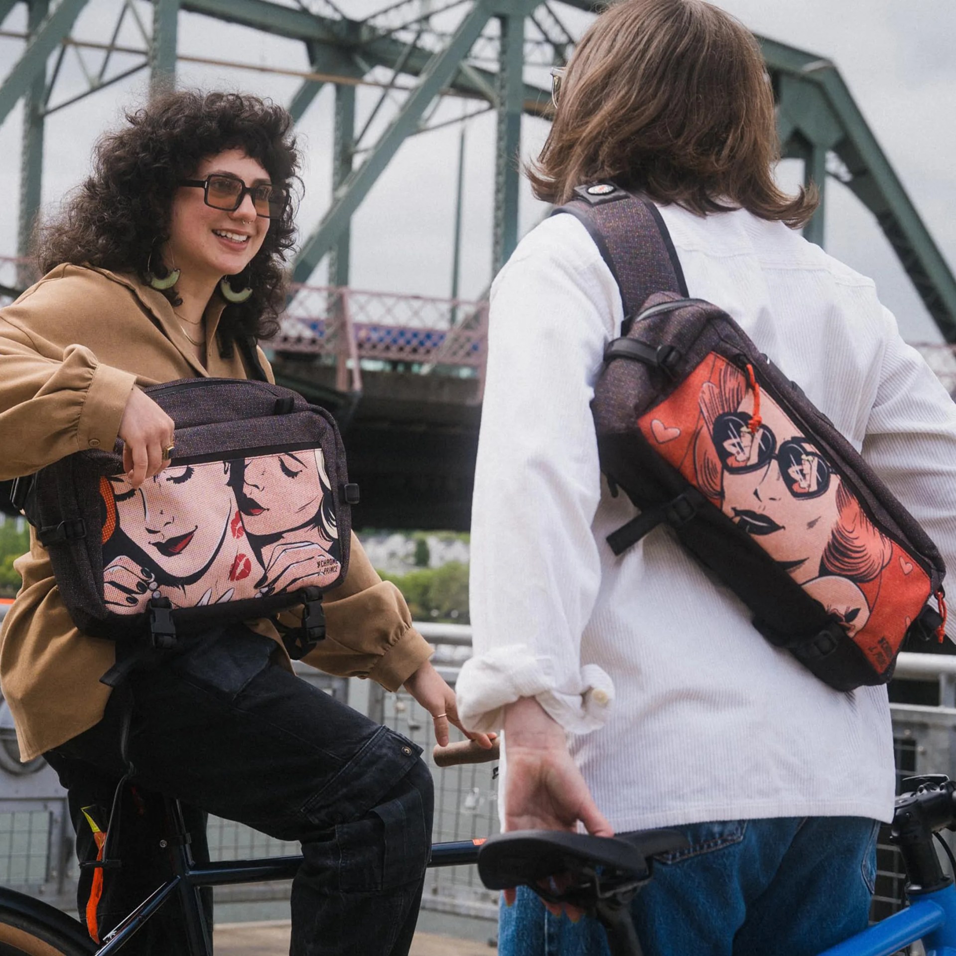 Two people with backpacks featuring pop art designs, one sitting on a bike and the other standing next to it under a metal bridge.
