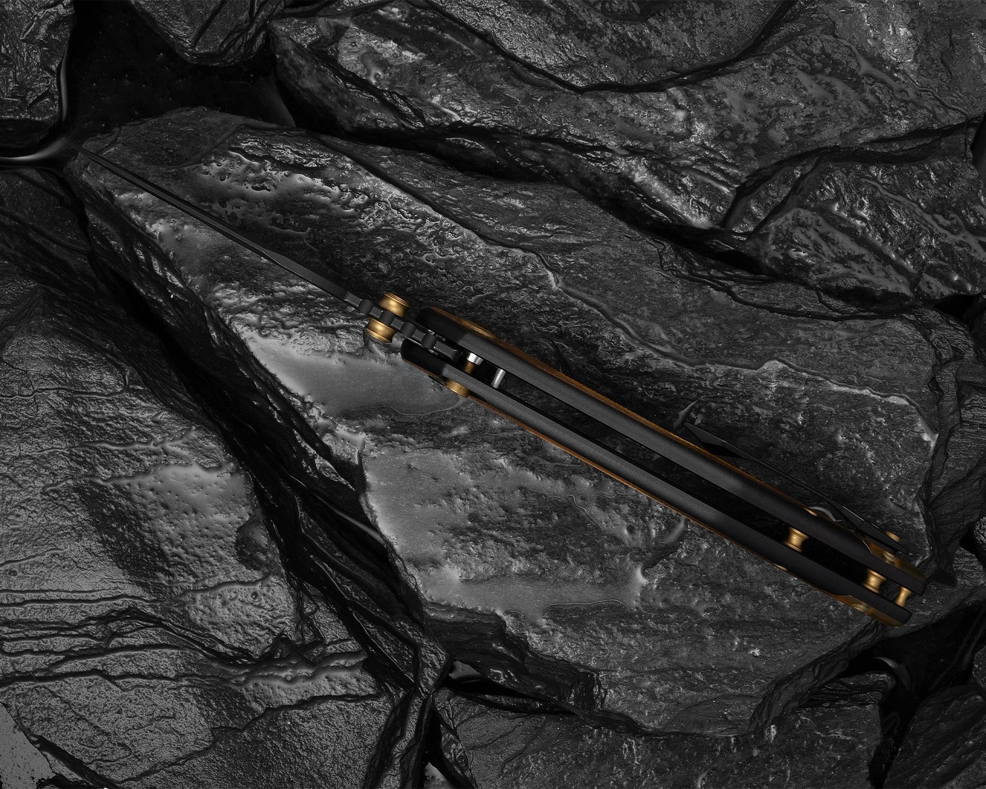 Close-up of a black folding knife with brass pivot on textured dark stone surface.