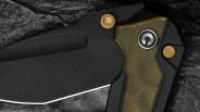 Close-up of a black folding knife blade with olive green textured handle and gold pivot screws on a dark textured surface.