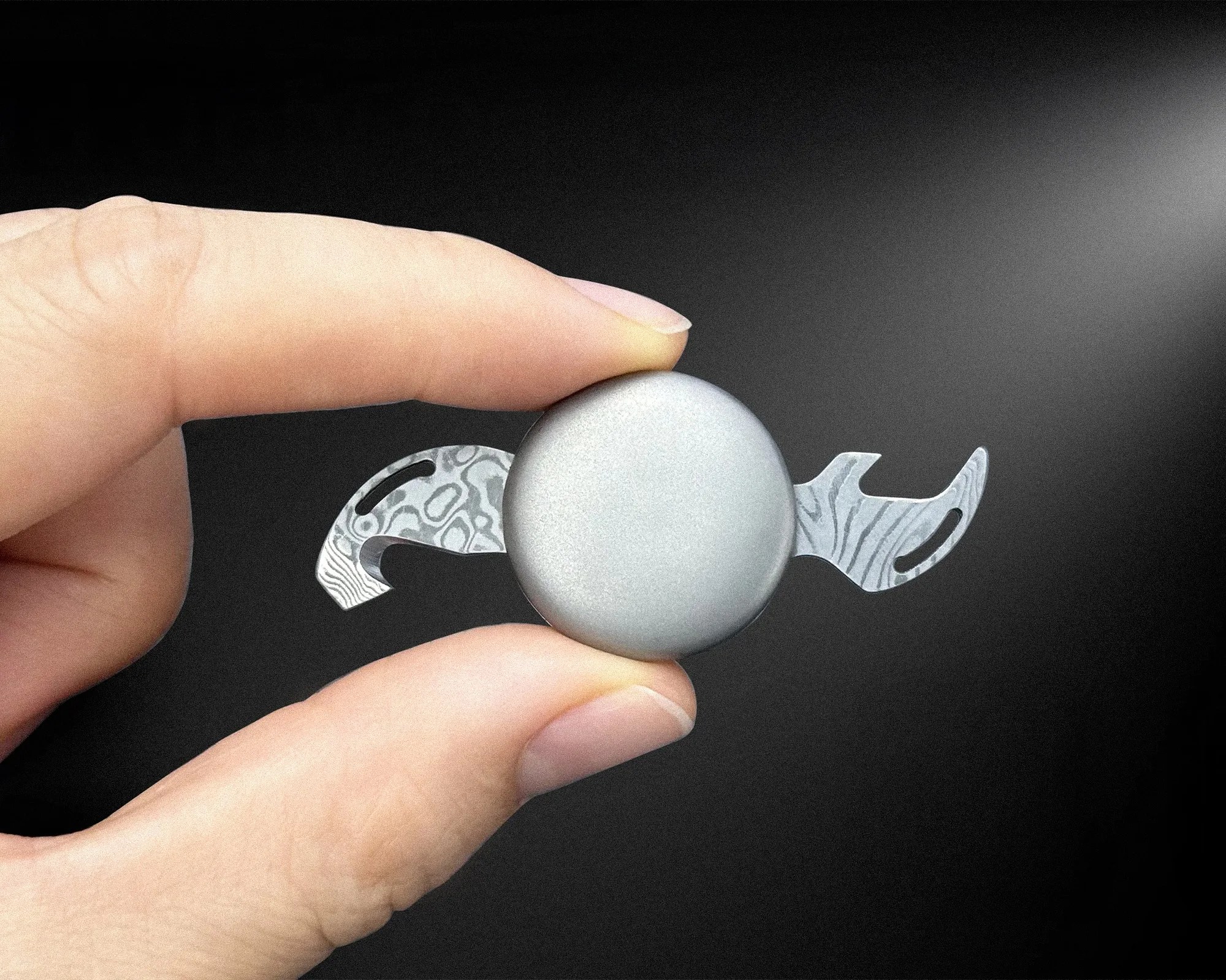 Hand holding a small metallic fidget spinner with patterned wings against a black background.