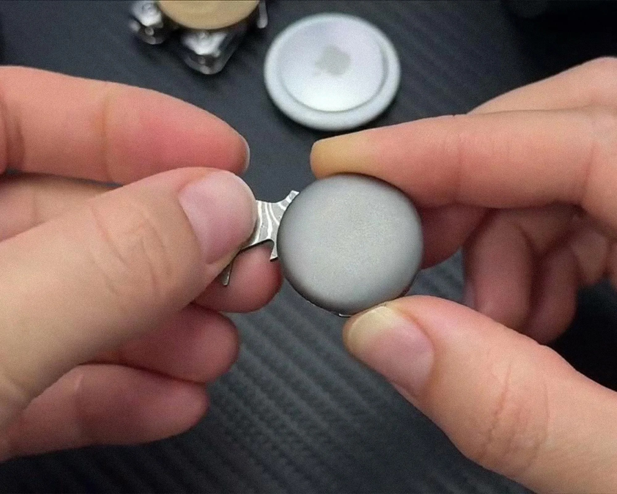 Hands holding a silver Apple AirTag with a small metal tool on a dark textured surface.