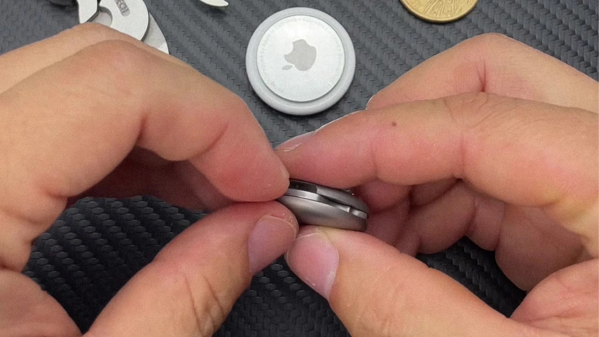 Hands holding and opening a silver Apple AirTag on a textured black surface with other AirTags and a coin nearby.