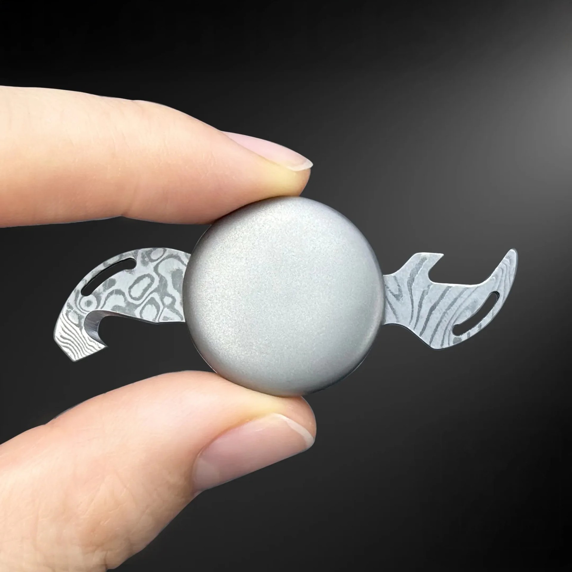 Fingers holding a small, round, silver metallic object with patterned extensions on a black background.