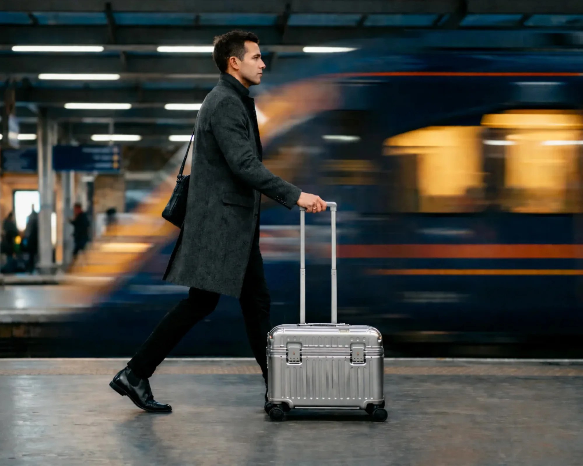 Man in a dark gray coat walking with a silver rolling suitcase at a train station with a blurred train in the background.