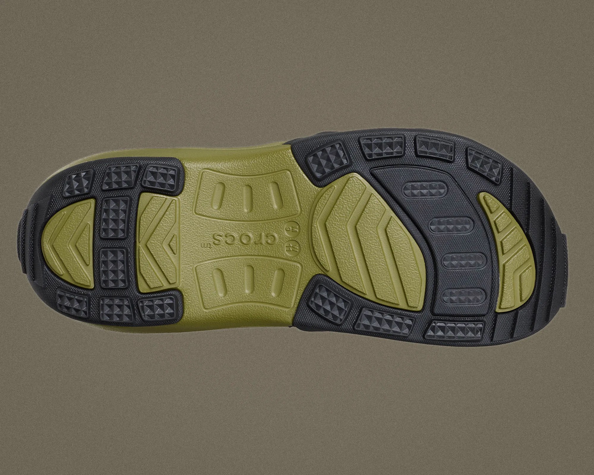 Olive green and black textured rubber sole of a Crocs shoe on a gray background.